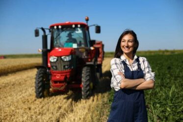 A woman in overalls standing in front of a tractor in a field. A Woman In Overalls Standing In Front Of A Tractor In A Field.