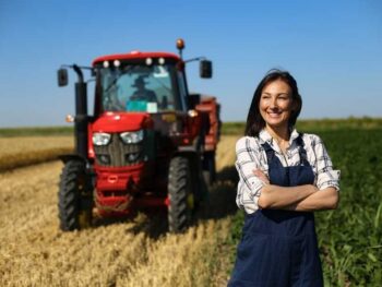 A Woman In Overalls Standing In Front Of A Tractor In A Field.