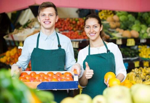 Two people standing in front of a fruit and vegetable stand with thumbs up. Two People Standing In Front Of A Fruit And Vegetable Stand With Thumbs Up.